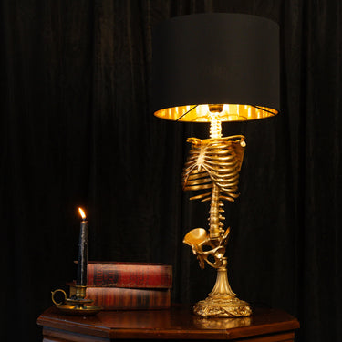 A skeleton decorative table lamp with a gold-toned rib cage and skull base, lit with a black drum shade and warm yellow interior light, set on a wooden table against a dark curtain backdrop. A candle and books sit beside it.