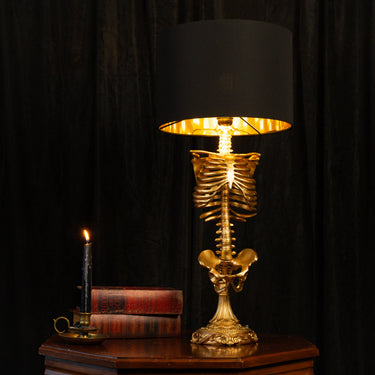 A decorative skeleton table lamp with a gold-toned skeleton rib cage and spine base, lit from within with a black drum shade and gold interior, set on a wooden table against a dark curtain backdrop.