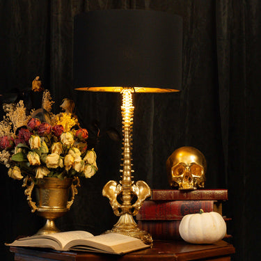 A decorative skeleton table lamp with a black drum shade and an ornate gold-toned stem and base, styled on a wooden surface beside a vase of flowers, an open book, stacked books, a gold skull, and a small white pumpkin against a dark curtain background.