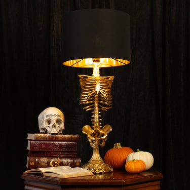 A skeleton-themed table lamp with a gold rib cage and spine base, featuring a black drum shade with a gold inner lining, displayed on a wooden table next to books, a skull, and small pumpkins against a black curtain background.