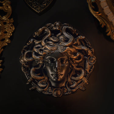 A circular wall-mounted sculpture of Medusa with snakes for hair, rendered in a dark metallic finish with warm gold highlights, set against a very dark background.