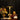 A decorative skeleton table lamp with a black drum shade and an ornate gold-toned stem and base, styled on a wooden surface beside a vase of flowers, an open book, stacked books, a gold skull, and a small white pumpkin against a dark curtain background.