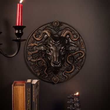 A circular resin gothic  wall plaque featuring a detailed ram's head surrounded by coiled snakes, styled on a dark wall with a candle holder, books, and another candle.