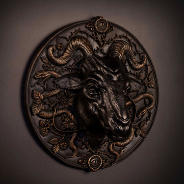An ornate circular wall sculpture featuring a detailed ram head in dark metallic finish, surrounded by coiled snakes, fruits, and leafy vines on a dark gray wall.